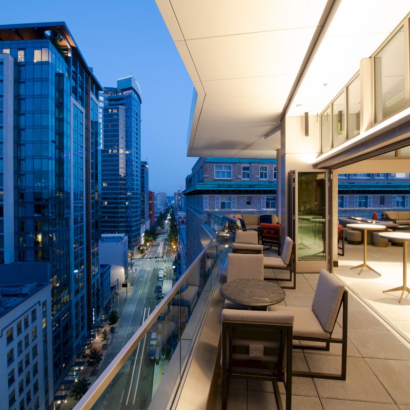 The image shows a cityscape at dusk from a high balcony with modern buildings, chairs, tables, and a glass railing overlooking the street below.