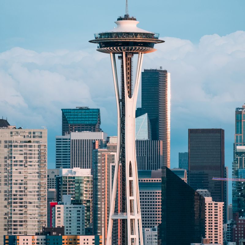 The image shows the iconic Space Needle in Seattle, Washington, with a backdrop of downtown skyscrapers and a partly cloudy sky.