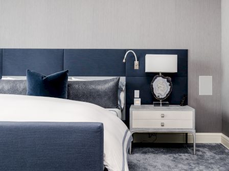 A modern bedroom with a blue and white color scheme, featuring a bed, pillows, a bedside table with a lamp, and an elegant wall-mounted light fixture.