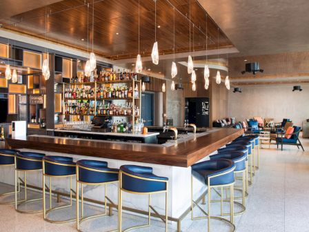 A modern bar with blue chairs, hanging lights, and a well-stocked drink selection, located in a spacious and stylish setting.