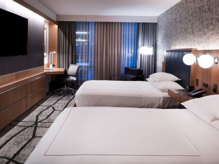 A modern hotel room with two beds, a desk with a chair, a wall-mounted TV, and patterned wallpaper, illuminated by soft, ambient lighting.