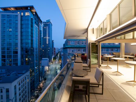The image shows a modern rooftop terrace overlooking a cityscape with tall buildings. The terrace has tables, chairs, and indoor-outdoor seating.