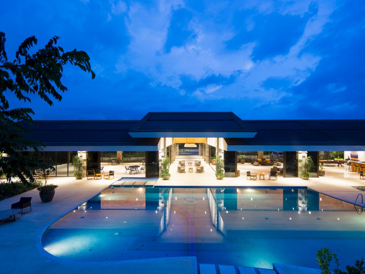A modern building with an illuminated pool in front during dusk, surrounded by open seating areas and under a partly cloudy sky.