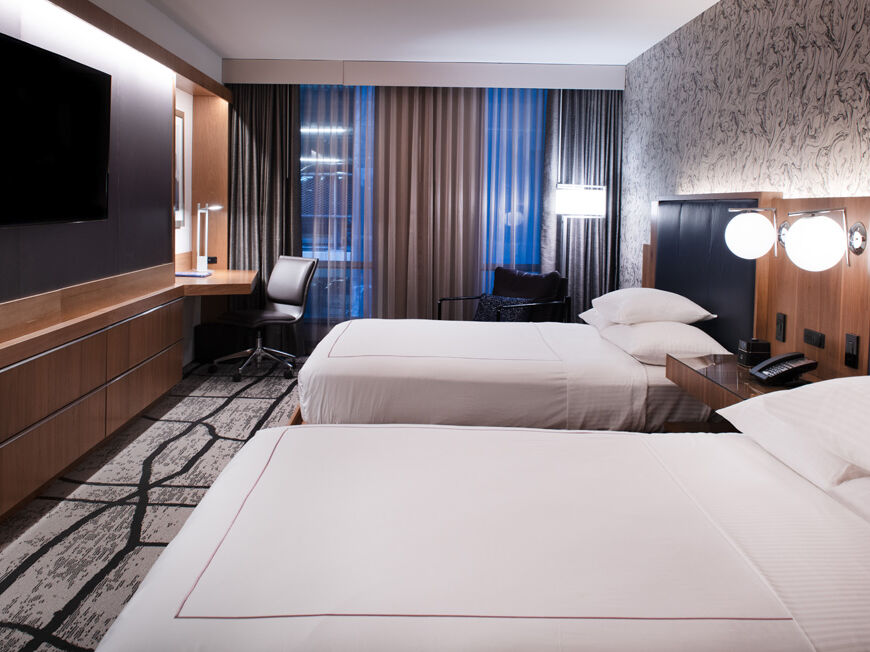 A modern hotel room with two double beds, a wall-mounted TV, work desk with chair, and a large window with curtains, all well-lit.