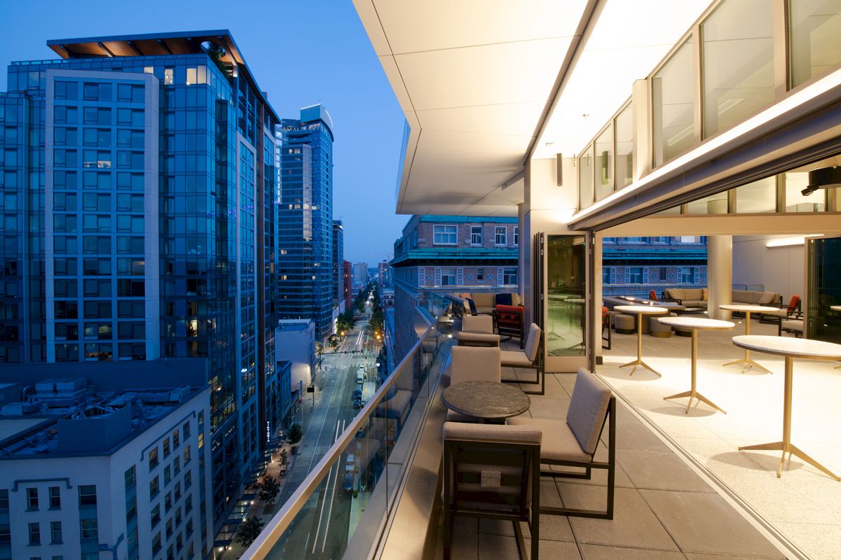 A modern balcony with tables and chairs overlooks a city street lined with tall buildings, illuminated during twilight hours, creating a serene atmosphere.