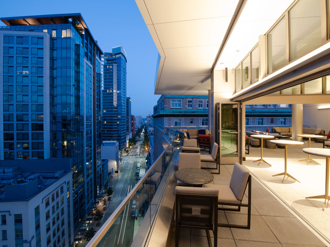A modern balcony with tables and chairs overlooks a city street lined with tall buildings, illuminated during twilight hours, creating a serene atmosphere.