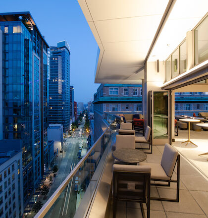 An outdoor deck with tables and chairs overlooks a cityscape with tall buildings at dusk. The deck has large glass doors and modern furniture.