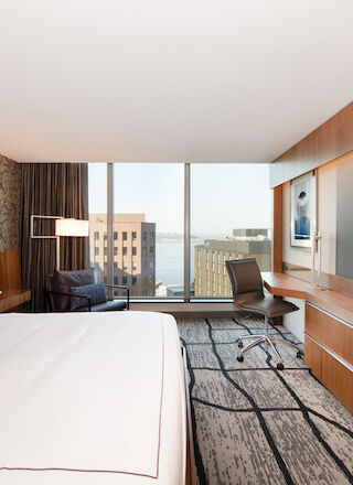 The image shows a modern hotel room with a large bed, a desk with a chair, a flat-screen TV, and a view of the city and waterfront.