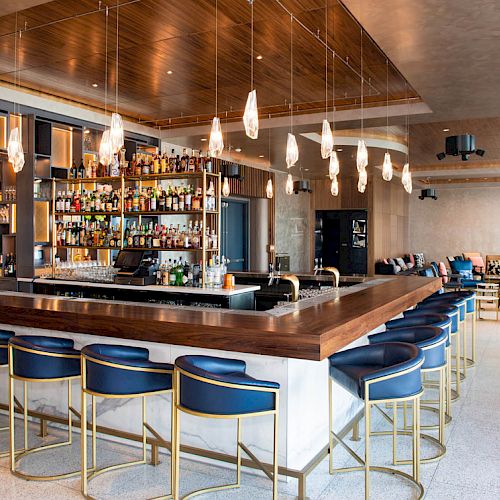 A modern bar interior featuring a central bar island with stools, hanging lights, and shelves stocked with various bottles. Lounge seating is in the background.