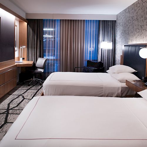 A modern hotel room with two beds, a wall-mounted TV, a desk with a chair, and contemporary decor including patterned wallpaper and carpet.