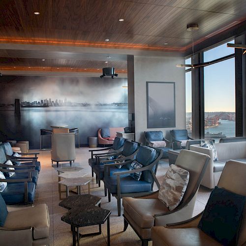 A modern lounge with numerous chairs, a city mural, large windows offering a scenic view of a harbor, and a Ferris wheel in the distance.