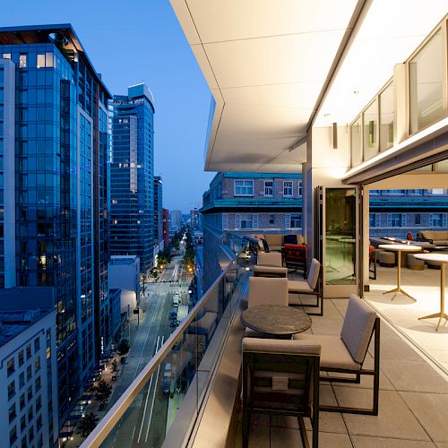 A modern urban balcony during evening with city buildings in the background. It features tables, chairs, and a view of a lit-up street.