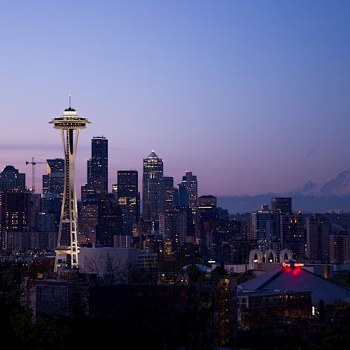 A cityscape of Seattle at dusk featuring the iconic Space Needle, various downtown buildings, and Mount Rainier in the distance.