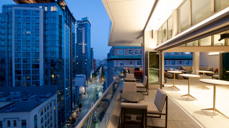 A modern rooftop terrace with tables and chairs, overlooking a city street lined with tall buildings at dusk, offering a stunning urban view.