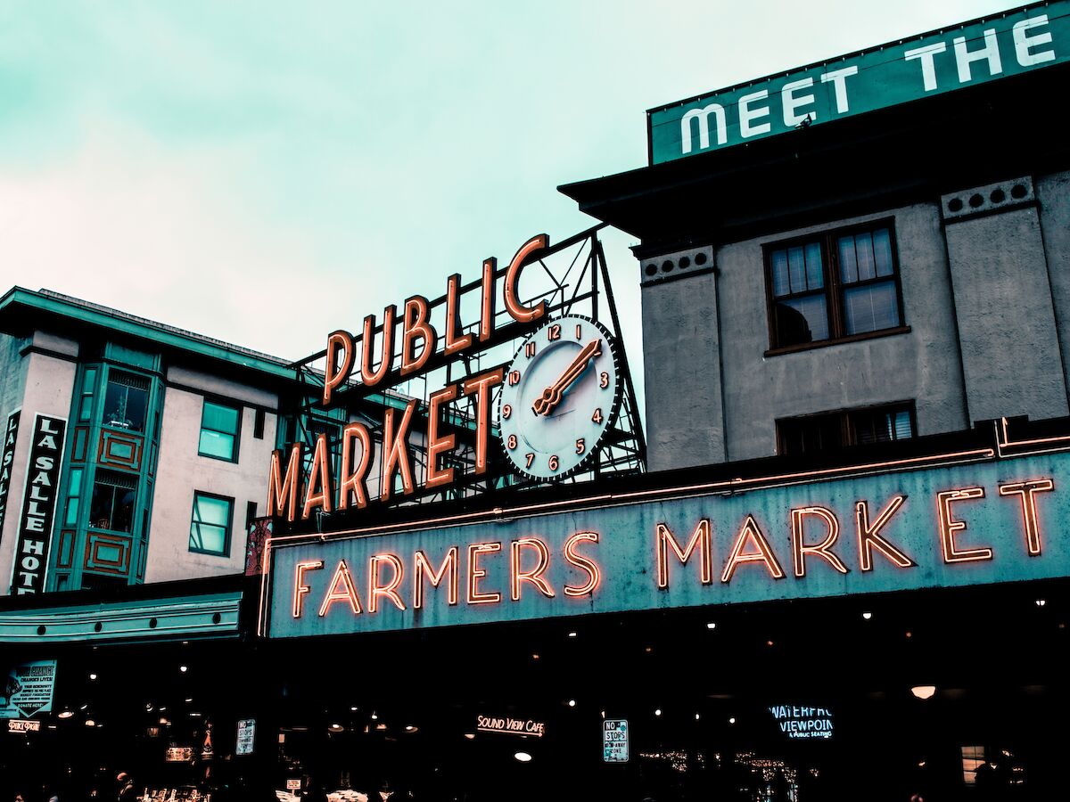 The image shows a neon sign for a "Public Market" and "Farmers Market" with a clock, in front of buildings. A sign partially reads "Meet the."