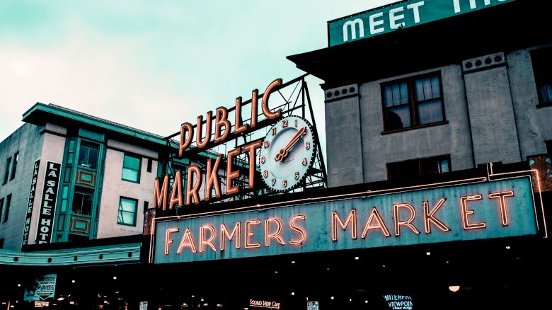 The image shows a neon sign for a "Public Market" and "Farmers Market" with a clock, in front of buildings. A sign partially reads "Meet the."