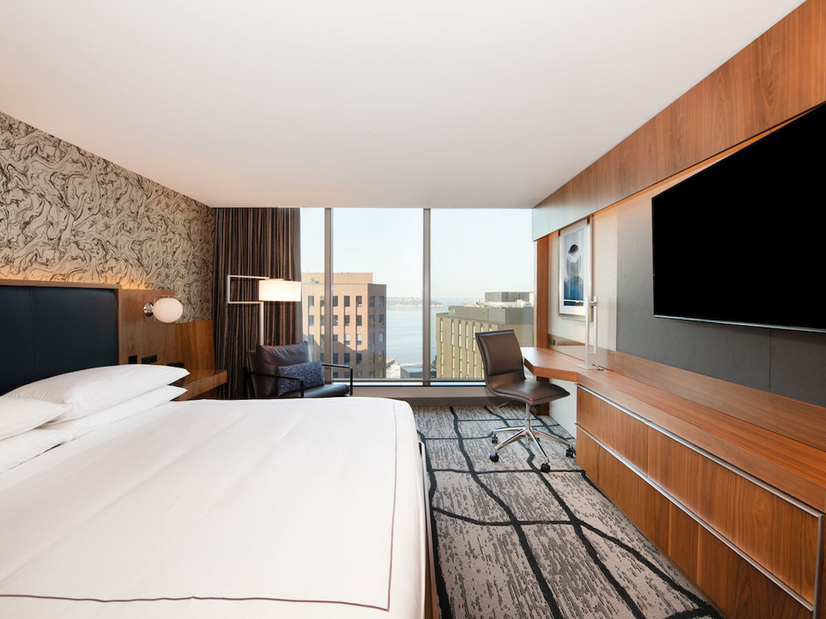 A modern hotel room features a large bed, a desk with a chair, a flat-screen TV, and a window with a view of city buildings and water.