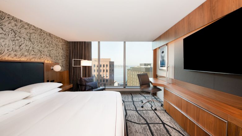 A modern hotel room features a large bed, a desk with a chair, a flat-screen TV, and a window with a view of city buildings and water.