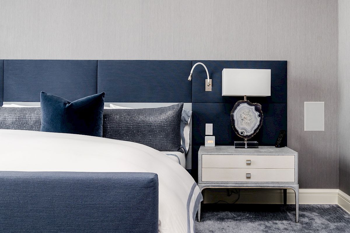 A modern bedroom features a blue upholstered headboard, white bedding, and a nightstand with a lamp and decorative item, on a grey-carpeted floor.