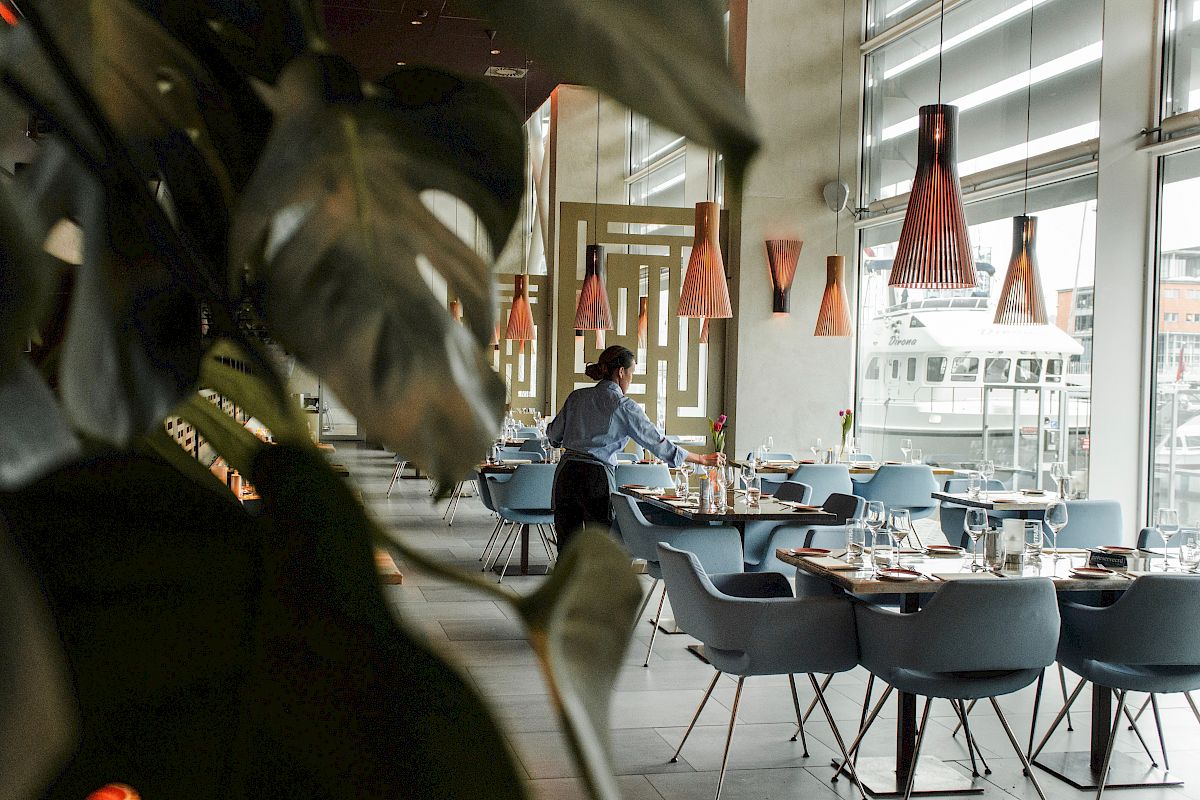 A man arranges tables in a modern restaurant with stylish lighting and large windows, while plants partially obstruct the view.